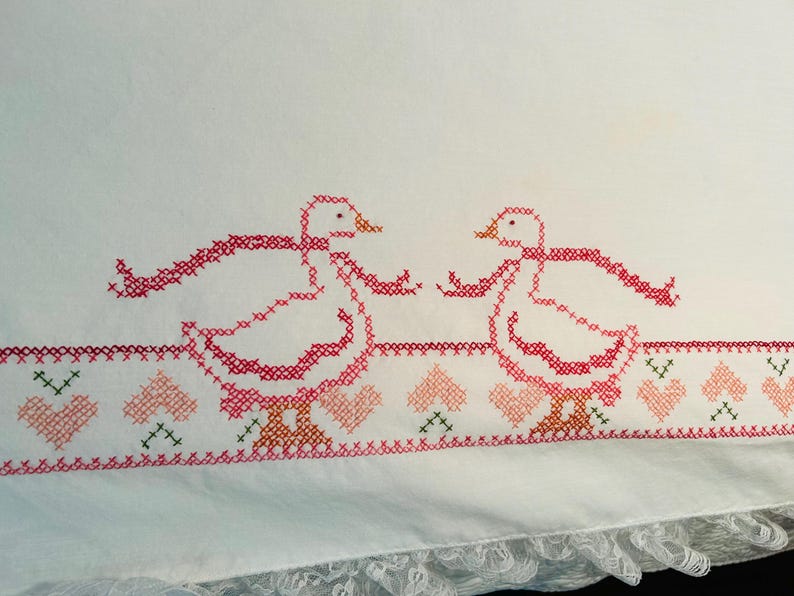 Puede incluir: Tela blanca con patos bordados a punto de cruz en rojo y rosa, cada uno con una cinta. Un borde de corazones y cruces en rosa, verde y rojo adorna el dise&ntilde;o. El borde inferior tiene un ribete de encaje.