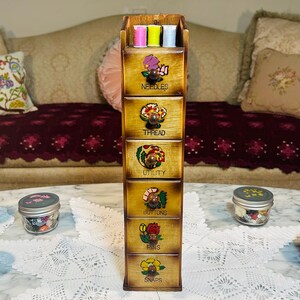 May include: A tall, vertical wooden storage cabinet with six drawers, each featuring a small round knob and a painted floral design. The drawers are labeled with the words NEEDLES, THREAD, UTILITY, BUTTONS, PINS, and SNAPS. Three spools of thread sit on top.