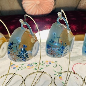 May include: Four decorative teacups with handles, displayed on gold-colored stands. The cups feature a design with blue floral patterns, brown and yellow accents, and a cross motif. The cups are displayed on a white tablecloth.