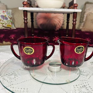 May include: A pair of deep red glass vessels, including a sugar bowl with two handles and a creamer pitcher with a spout and single handle. Both pieces feature a circular yellow sticker with the text Royal Ruby Anchorglass printed in black.