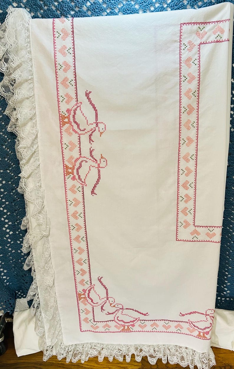 Puede incluir: Mantel de lino blanco con bordado de punto de cruz rosa. El bordado presenta un borde de corazones y gansos, con un ribete de encaje en el borde. El dise&ntilde;o es de estilo cl&aacute;sico y vintage.