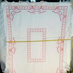 Puede incluir: Mantel blanco con bordado de punto de cruz rosa. El bordado presenta un patr&oacute;n repetitivo de corazones y p&aacute;jaros estilizados. Los bordes est&aacute;n adornados con un encaje delicado. Una cinta m&eacute;trica amarilla se extiende sobre el mantel.