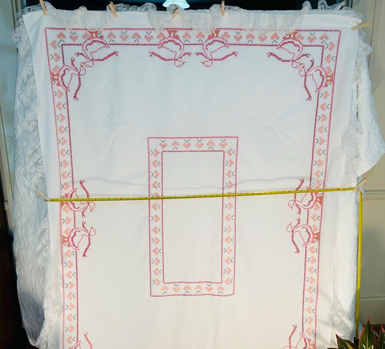 Puede incluir: Mantel blanco con bordado de punto de cruz rosa. El bordado presenta un patr&oacute;n repetitivo de corazones y p&aacute;jaros estilizados. Los bordes est&aacute;n adornados con un encaje delicado. Una cinta m&eacute;trica amarilla se extiende sobre el mantel.
