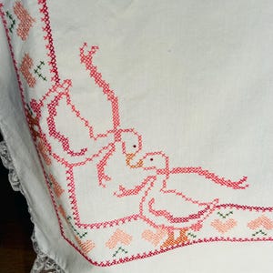 Puede incluir: Tela blanca bordada con un dise&ntilde;o de punto de cruz rojo y rosa. El dise&ntilde;o presenta dos gansos, corazones y detalles florales. El borde est&aacute; adornado con un delicado encaje blanco.