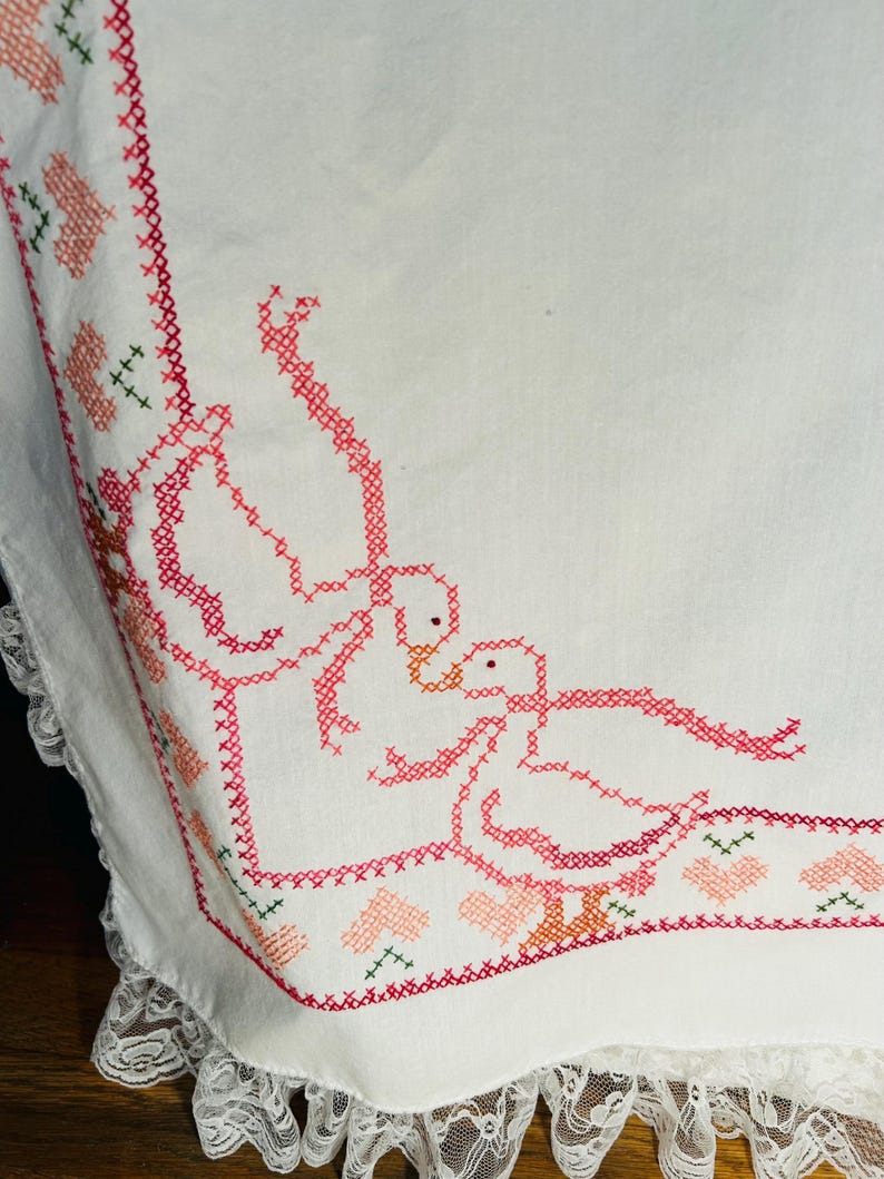 Puede incluir: Tela blanca bordada con un dise&ntilde;o de punto de cruz rojo y rosa. El dise&ntilde;o presenta dos gansos, corazones y detalles florales. El borde est&aacute; adornado con un delicado encaje blanco.