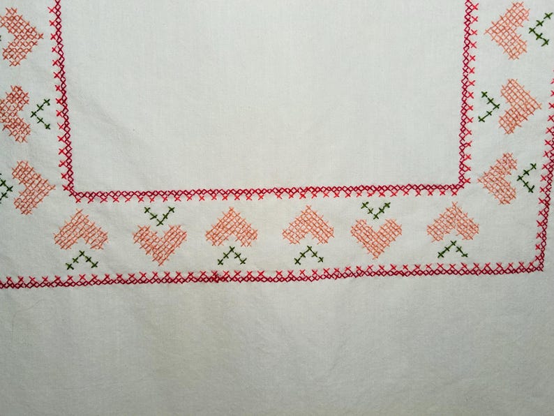 Puede incluir: Tela blanca con un borde de punto de cruz rojo y verde. El borde presenta corazones de color melocot&oacute;n y detalles verdes de punto de cruz. Las esquinas tienen un dise&ntilde;o similar, creando un patr&oacute;n cohesivo.