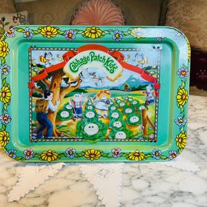 May include: A rectangular, vintage metal tray with a teal background and a colorful floral border. The tray features a central illustration of Cabbage Patch Kids in a field, with the logo at the top. The tray is likely a collectible item.