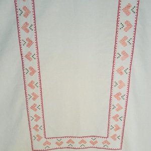 Puede incluir: Tela blanca con un borde decorativo que presenta un patr&oacute;n repetitivo de corazones rosas y detalles de punto de cruz verdes. El borde est&aacute; delineado con una puntada roja. El dise&ntilde;o es sim&eacute;trico y recorre los bordes de la tela.