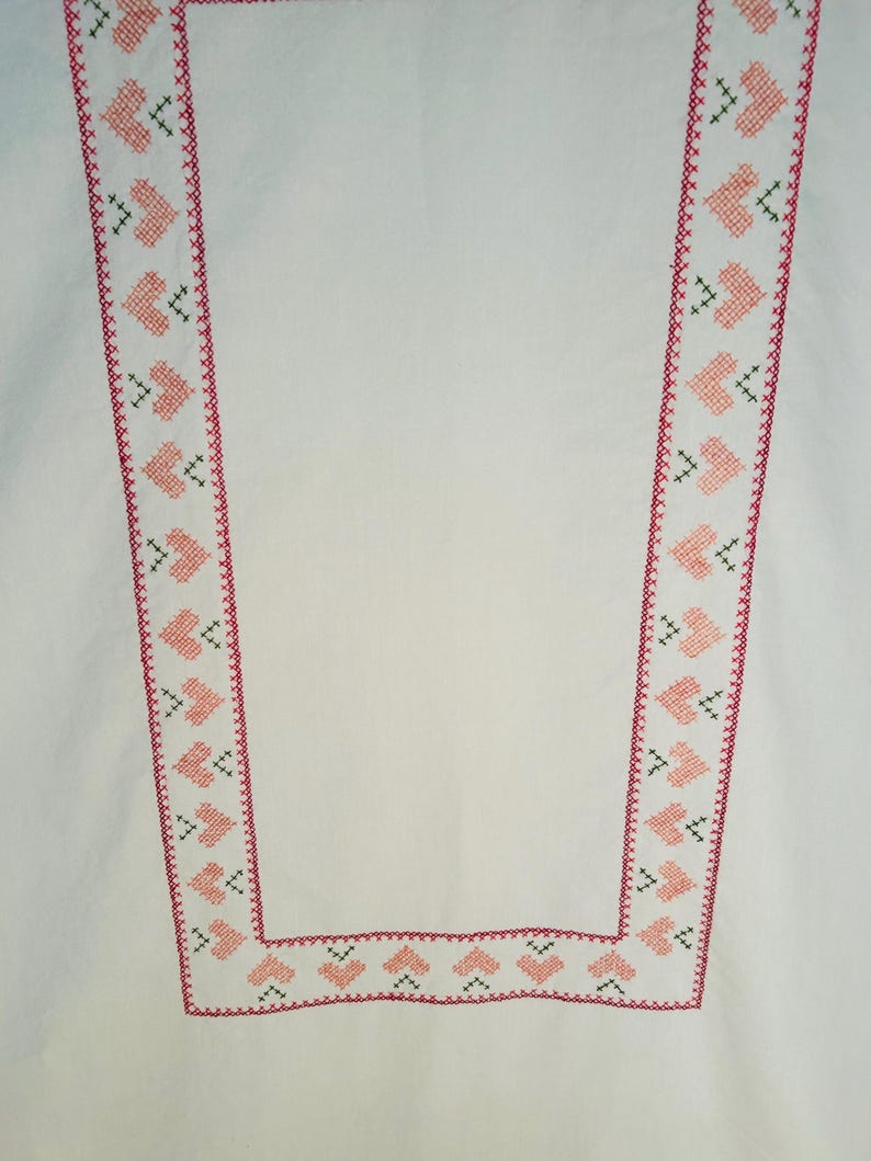 Puede incluir: Tela blanca con un borde decorativo que presenta un patr&oacute;n repetitivo de corazones rosas y detalles de punto de cruz verdes. El borde est&aacute; delineado con una puntada roja. El dise&ntilde;o es sim&eacute;trico y recorre los bordes de la tela.