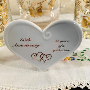 May include: A white heart-shaped ceramic dish with gold text that reads "50th Anniversary" and "60 years of a golden love." Two gold intertwined hearts are below the text. Clear glass mugs with gold "Anniversary" text are in the background.