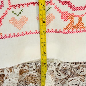 Puede incluir: Tela blanca con bordado de punto de cruz en rojo, rosa y naranja, con corazones y motivos florales. La tela tiene un ribete de encaje en la parte inferior. La medida es de 175 cm.