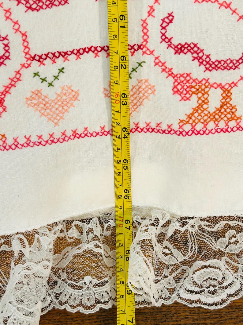 Puede incluir: Tela blanca con bordado de punto de cruz en rojo, rosa y naranja, con corazones y motivos florales. La tela tiene un ribete de encaje en la parte inferior. La medida es de 175 cm.