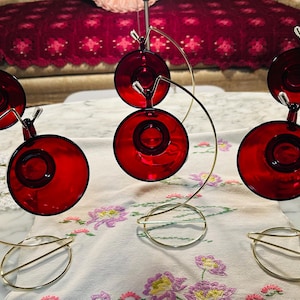 May include: Six red glass candle holders with a circular design are displayed on a white tablecloth with floral embroidery. Each candle holder is suspended by a gold-colored metal stand. The background includes a burgundy sofa.