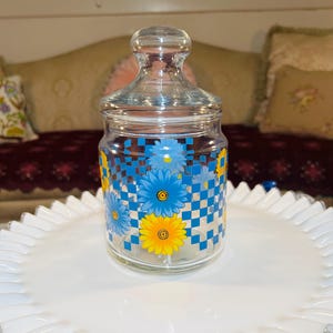 May include: A clear glass canister with a matching lid featuring a knob handle. The exterior surface is decorated with a repeating blue and white checkerboard pattern interspersed with vibrant yellow and blue daisy flower illustrations.