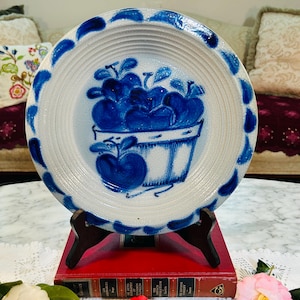 May include: A white ceramic plate with a blue apple basket design. The plate has a ridged center and a blue dotted border. The plate is displayed on a wooden stand, with a red book beneath it.