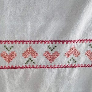 Puede incluir: Tela blanca con un borde de corazones bordados a punto de cruz y costuras decorativas. Los corazones son rosas y las costuras son rojas y verdes. La tela tiene un aspecto texturizado.
