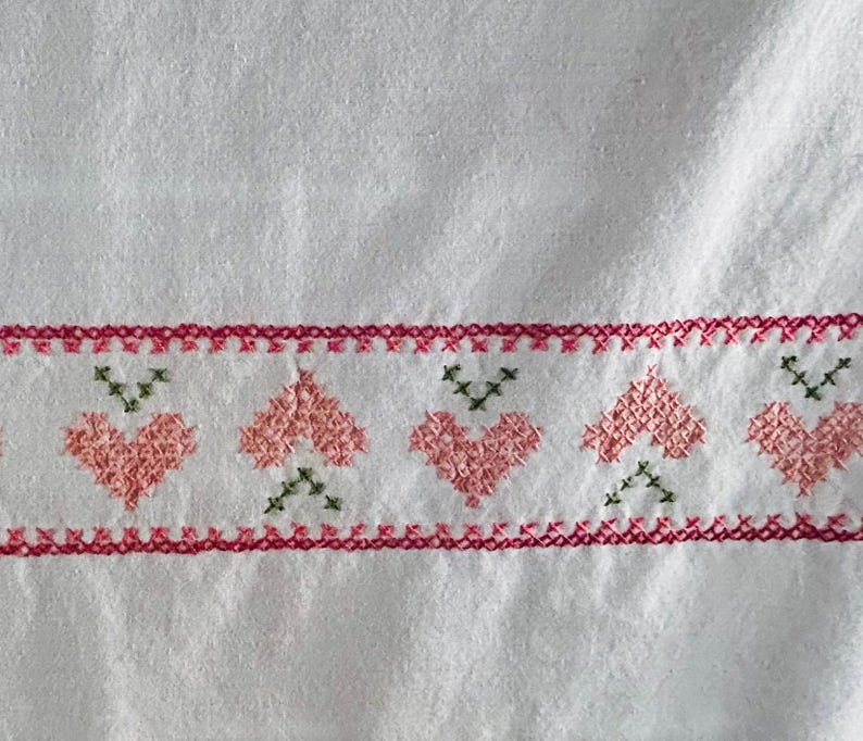 Puede incluir: Tela blanca con un borde de corazones bordados a punto de cruz y costuras decorativas. Los corazones son rosas y las costuras son rojas y verdes. La tela tiene un aspecto texturizado.
