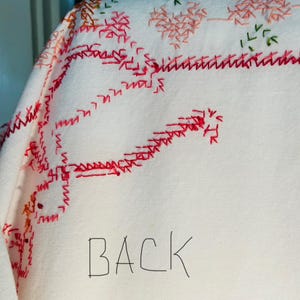 Puede incluir: Primer plano de un textil blanco con patrones florales y geom&eacute;tricos bordados en rojo, naranja y verde. La palabra "BACK" est&aacute; escrita a mano con un rotulador negro sobre la tela.