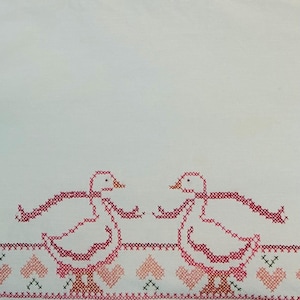 Puede incluir: Tela blanca con bordado de punto de cruz. El dise&ntilde;o presenta dos gansos rosas enfrentados, con un borde de corazones rosas y cruces negras. Los gansos tienen las alas extendidas, creando un dise&ntilde;o artesanal.