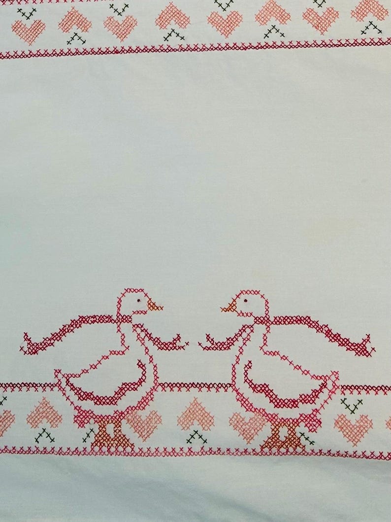 Puede incluir: Tela blanca con bordado de punto de cruz. El dise&ntilde;o presenta dos gansos rosas enfrentados, con un borde de corazones rosas y cruces negras. Los gansos tienen las alas extendidas, creando un dise&ntilde;o artesanal.