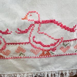 Puede incluir: Tela blanca con un dise&ntilde;o de punto de cruz que presenta un pato rosa con un pico naranja y un lazo rosa. El borde inferior tiene un borde de corazones y flores rosas y un ribete de encaje blanco.