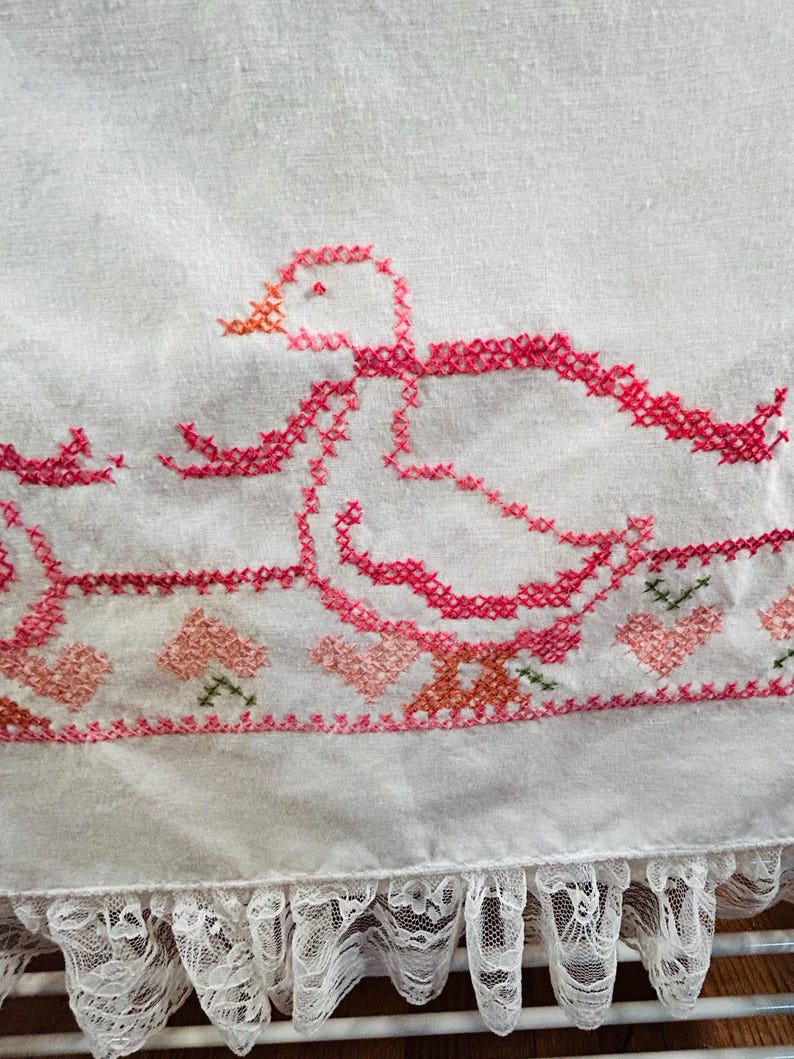 Puede incluir: Tela blanca con un dise&ntilde;o de punto de cruz que presenta un pato rosa con un pico naranja y un lazo rosa. El borde inferior tiene un borde de corazones y flores rosas y un ribete de encaje blanco.