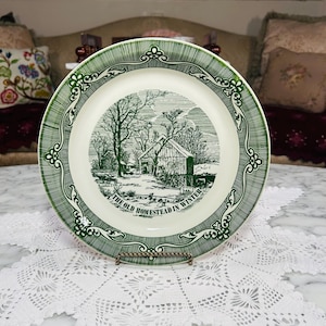 May include: A round ceramic plate with a cream-colored center and a dark green border. The center features a detailed black and white illustration of a barn in a snowy landscape with the text, THE OLD HOMESTEAD IN WINTER, printed below.