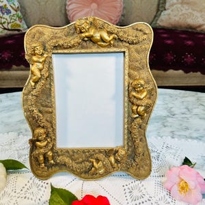 May include: Ornate gold-colored picture frame with cherub accents. The frame has a rectangular opening and is decorated with multiple cherub figures and floral details. The frame is sitting on a white lace tablecloth.