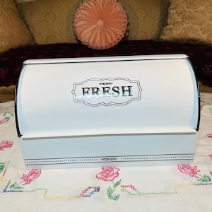 May include: White metal bread box with a curved lid and black trim. The word "FRESH" is displayed in a decorative frame on the front. A dotted line with leaf accents decorates the bottom of the box. The box is on a floral patterned tablecloth.