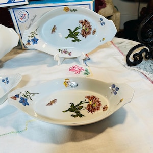 May include: Two white oval serving dishes with floral and butterfly designs. Each dish features a central flower with brown and green petals, surrounded by butterflies and colorful flowers. The dishes have handles on each side.