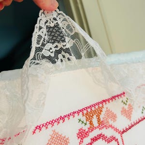 Puede incluir: Primer plano de un textil blanco bordado con un dise&ntilde;o floral en tonos rosa, naranja y verde. El borde est&aacute; adornado con un delicado encaje blanco. El textil est&aacute; sostenido por una mano.