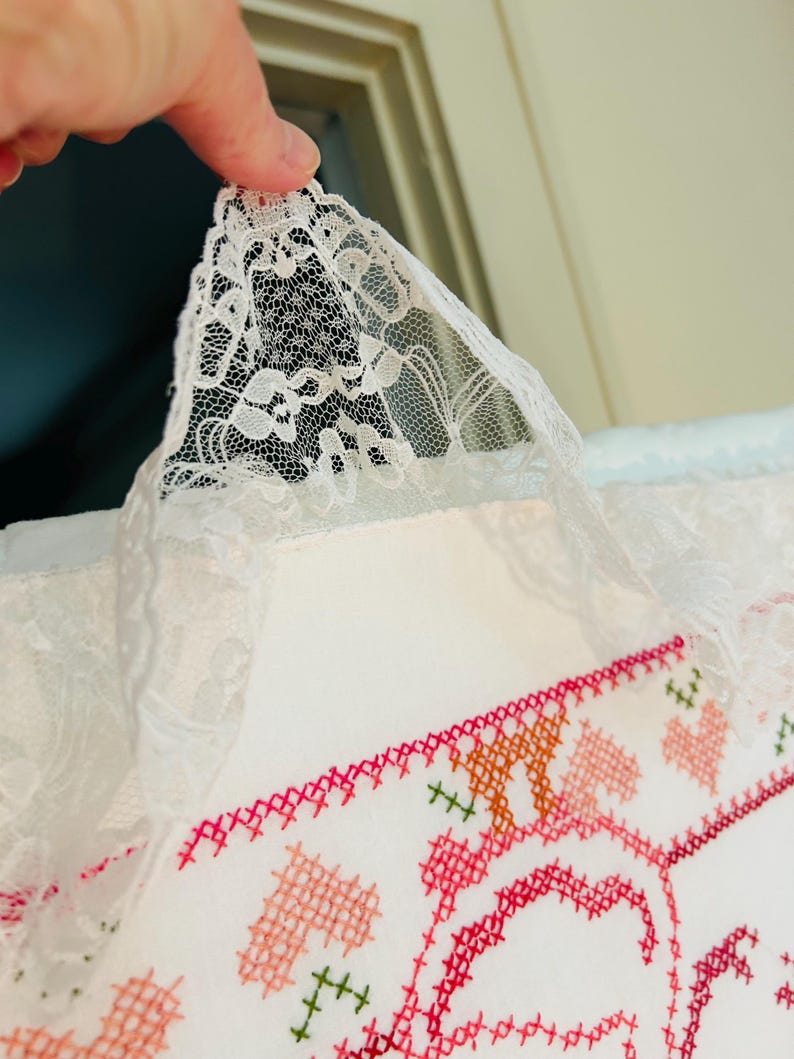 Puede incluir: Primer plano de un textil blanco bordado con un dise&ntilde;o floral en tonos rosa, naranja y verde. El borde est&aacute; adornado con un delicado encaje blanco. El textil est&aacute; sostenido por una mano.