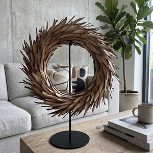 May include: A circular wreath composed of numerous pointed, dried brown leaves arranged in a radial pattern, mounted on a slender black metal stand with a circular base. The leaves have a textured, organic appearance with sharp, tapered tips.