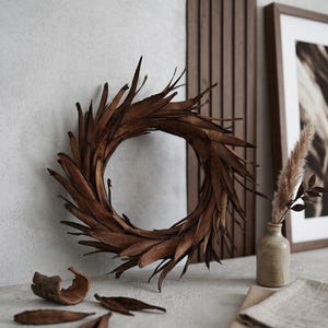 Pode incluir: Uma coroa circular de folhas castanhas. A coroa é exibida numa superfície cinza claro, com um pequeno vaso de plantas secas e uma imagem emoldurada no fundo. Folhas e casca adicionais estão espalhadas ao redor da coroa.