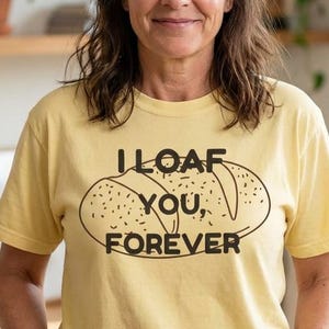 May include: A light yellow t-shirt with the text "I LOAF YOU, FOREVER" and a bread loaf graphic. The shirt is worn by a person with dark hair and blue jeans. The background includes a wooden shelf with plants.