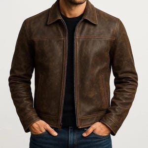 Puede incluir: Chaqueta de cuero marrón con cuello y cierre de cremallera. La chaqueta tiene dos bolsillos delanteros y se usa sobre un suéter negro y jeans azules.