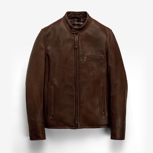 Puede incluir: Chaqueta de cuero marrón con cremallera, un pequeño bolsillo en el pecho y dos bolsillos laterales. La chaqueta tiene un cuello alzado y un forro a cuadros visible en el cuello. El cuero tiene un aspecto ligeramente desgastado.