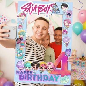 May include: A light blue photo booth prop with the text "SAJA BOYS" and "HAPPY BIRTHDAY". The prop has cartoon character illustrations and the number "4". The photo booth is being held by a person taking a selfie with two other children.