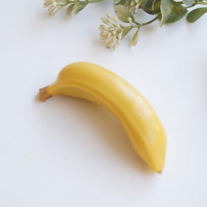 Może przedstawiać: Pojedynczy, gładki banan o jasnym, żółtym kolorze, o lekko zakrzywionym kształcie z małą, brązową końcówką łodygi. Banan jest ułożony poziomo, prezentując swój jednolity kolor oraz realistyczną, twardą teksturę odbijającą światło na powierzchni.