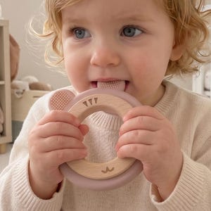 May include: A baby chews on a wooden and silicone teething ring. The ring has a bear design and is a pale pink and natural wood colour. The baby wears a cream-coloured jumper and sits on a fluffy, light-coloured rug.