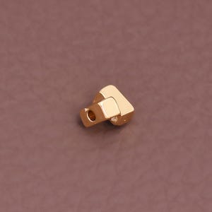 May include: A small, gold-colored metal object with a geometric shape. It has a square body with a cylindrical hole on one side and a curved, angled extension. The object is set against a solid, brown background.