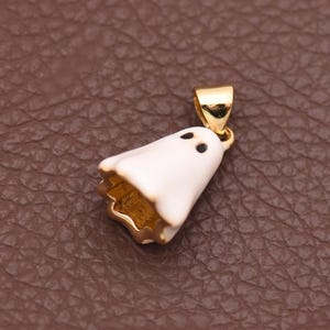 May include: A gold-toned metal pendant shaped like a ghost, featuring a smooth white enamel coating on the exterior and two small black dots representing eyes. The pendant includes a small gold-toned bail at the top for attachment to a chain.