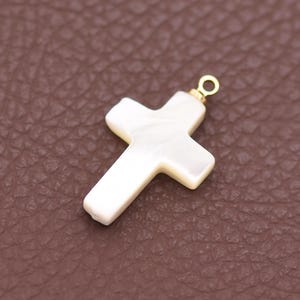 May include: A white cross pendant with a pearlescent finish and a small gold-colored loop at the top. The cross has a classic design and is likely made of a natural material. The pendant is shown against a brown background.