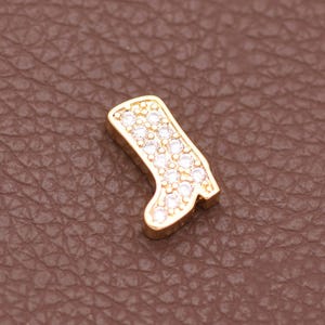 May include: A small gold-toned charm shaped like a boot, featuring a series of clear, sparkling stones embedded along the front surface. The boot charm has a defined heel and shaft, with a polished metallic finish surrounding the inlaid stones.