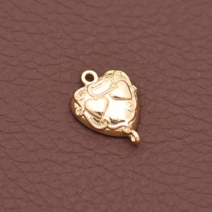 May include: A small gold-coloured metal charm in the shape of a heart. The surface features an embossed design with two smaller heart shapes and swirling lines. There is a small circular loop at the top and a smaller loop at the bottom for attachment.