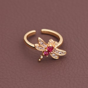 May include: A gold-colored adjustable ring featuring a dragonfly design. The dragonfly has pink and clear crystal accents. The ring is set against a brown background.