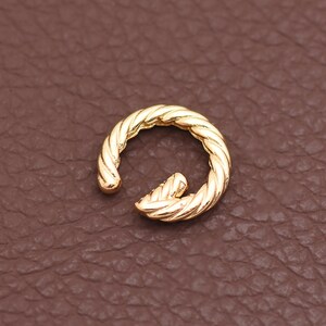 Puede incluir: Un pendiente tipo ear cuff de metal dorado con una textura de cuerda trenzada. La pieza circular está abierta, con extremos redondeados, y muestra un acabado pulido y reflectante. El diseño consiste en un patrón en espiral continuo.