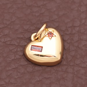 May include: A gold-colored heart-shaped pendant with a small red rectangular stone and a red star-shaped stone. The pendant has a loop at the top for a chain. The heart is smooth and shiny.