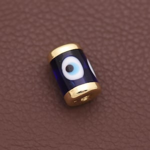 May include: A cylindrical bead featuring a deep blue glass body with a white and black eye motif. The bead is capped on both ends with polished gold-toned metal, and it has a central hole running through the length of the cylinder for threading.