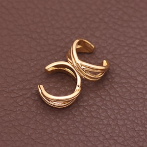 May include: A pair of gold-toned ear cuffs featuring a multi-band design. Each piece consists of three thin, parallel metal bands that curve into an open circular shape, creating a layered appearance with a polished, reflective finish.